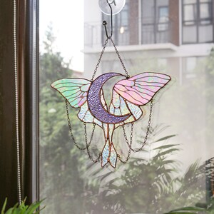 May include: A stained glass window hanging with a crescent moon and a moth design. The moon is purple and the moth has iridescent wings with pink, green, and blue hues.