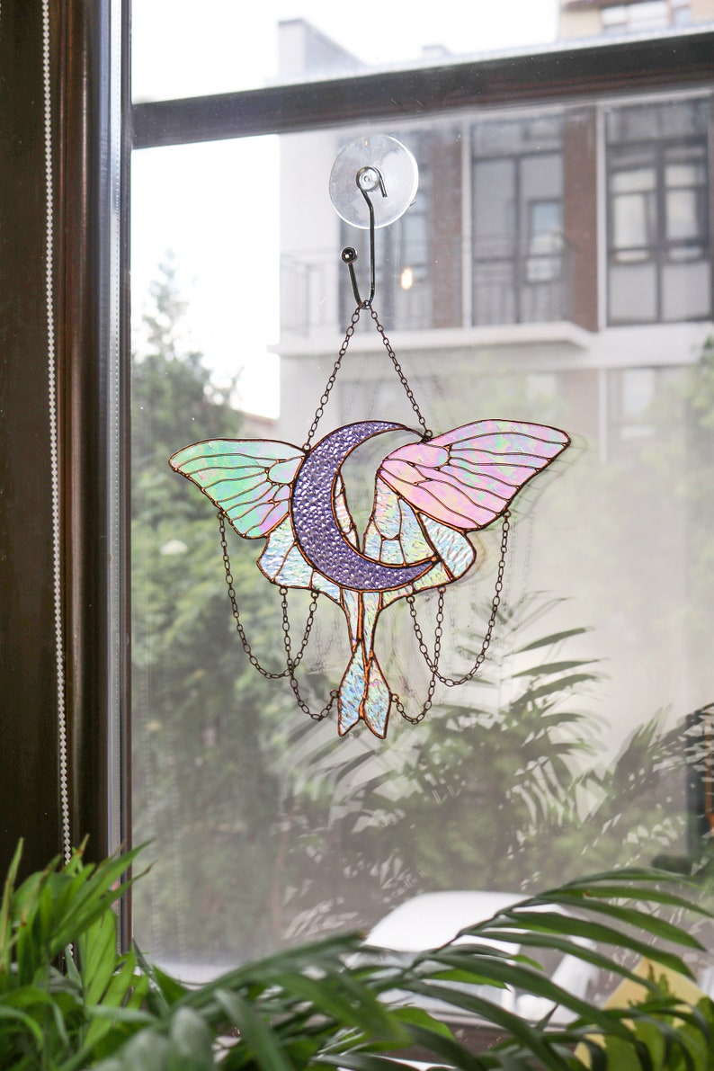 May include: A stained glass window hanging with a crescent moon and a moth design. The moon is purple and the moth has iridescent wings with pink, green, and blue hues.