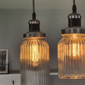 May include: Two clear glass pendant lights with a ribbed texture and a black metal frame. The lights are turned on and illuminate the room.