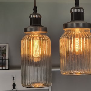 Könnte beinhalten: Zwei klare Glaspendelleuchten mit gerippten Texturen und warmweißen Glühbirnen. Die Leuchten haben eine silberne Metalloberfläche und hängen an einer Decke.