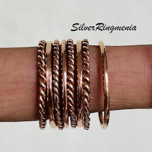 Puede incluir: Una colección de anillos de color cobre, algunos con un diseño de cuerda trenzada, se muestra en un dedo. Los anillos varían en grosor, con una banda fina y lisa al final. La marca "Silver Ringmenia" es visible.