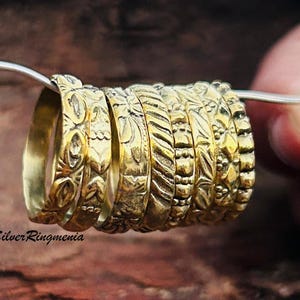 May include: A set of eight gold-toned stacking rings with different embossed patterns. The rings are stacked on top of each other and are being held by a hand.