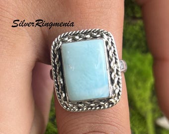 May include: A silver ring with a rectangular, pale blue gemstone. The stone is framed by a decorative, rope-style border. The ring is on a finger, with the text "Silver Ringmenia" visible.