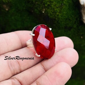 May include: A silver ring featuring a large, faceted, oval-shaped red gemstone. The gemstone is the focal point, with a brilliant, reflective surface. The ring's band is a simple, polished silver. The text "Silver Ringmenia" is visible on the image.