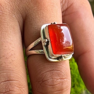 Peut inclure: Bague en argent ornée d'une grosse pierre précieuse rectangulaire polie, rouge orangé. La pierre est sertie dans un cadre carré avec des ornements décoratifs. La bague est portée sur un doigt, sur fond vert flou.
