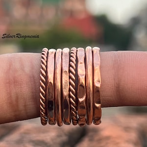Puede incluir: Un conjunto de anillos de color cobre, cada uno con una textura única, incluyendo diseños trenzados y martillados. Los anillos están apilados en un dedo, mostrando sus diferentes estilos y brillo metálico. Los anillos tienen un tono cálido, rojizo-marrón.