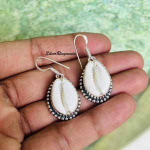 Puede incluir: Un par de pendientes de plata con colgantes de concha blanca. Los pendientes tienen un borde plateado oscuro detallado y un diseño de gancho simple. La imagen incluye el texto "Silver Ringmenia".