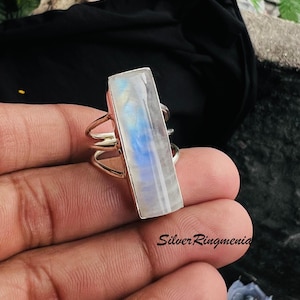 May include: A silver ring featuring a long, rectangular moonstone cabochon with a blue iridescent sheen. The band is constructed from multiple thin, parallel silver wires that converge at the sides. The text SilverRingmenia is visible.