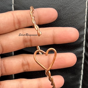 Puede incluir: Dos anillos de cobre. Un anillo es una banda retorcida. El otro anillo tiene forma de corazón con una base de banda retorcida. Los anillos se muestran sobre un fondo oscuro. Los anillos son hechos a mano y tienen un acabado brillante.