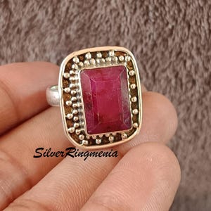 Può includere: Un anello d'argento con una grande gemma rettangolare rosso intenso. La gemma è incastonata in una cornice quadrata con un bordo strutturato e puntinato. La fascia dell'anello è semplice e argentata. Il testo "Silver Ringmenia" è visibile.