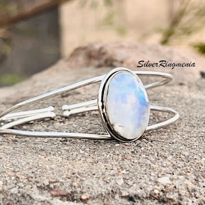 Peut inclure: Un bracelet manchette argenté avec une pierre de lune ovale et irisée. La pierre de lune présente des nuances de bleu et de blanc. Le bracelet a un design ouvert et simple. Le texte "Silver Ringmenia" est visible.
