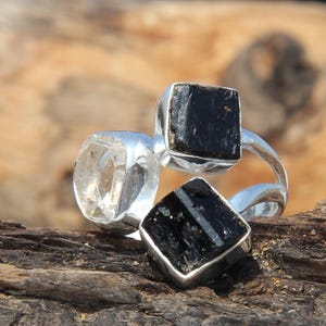 Puede incluir: Tres anillos de piedras preciosas. Un anillo presenta una piedra clara y facetada. Dos anillos tienen piedras negras cuadradas engastadas en bandas de plata. Los anillos se exhiben sobre un trozo de madera envejecida.