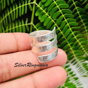 Puede incluir: Un anillo plateado envolvente con textura martillada. El anillo tiene un diseño en espiral con tres bandas que se envuelven alrededor del dedo. El texto "Silver Ringmenia" es visible en la parte inferior de la imagen.
