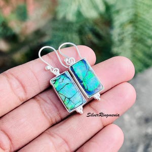 May include: A pair of rectangular silver earrings with iridescent blue and green stones. The stones have a cracked, mosaic-like pattern. The earrings are held by a hand, with the brand name "SilverRingmenia" visible.