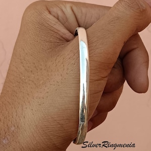 May include: A silver bangle bracelet with a smooth, polished finish. The bracelet is shown on a person's wrist.
