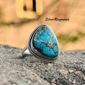 May include: A silver ring with a large, teardrop-shaped turquoise stone. The stone has a matrix of gold-colored veins. The ring's band is silver, and the stone is framed by a twisted rope design. The text "Silver Ringmenia" is visible.