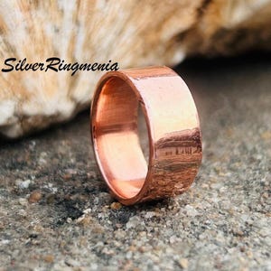 Op de afbeelding: Een brede koperen ring, geplaatst op een grijze, getextureerde ondergrond. Het oppervlak van de ring reflecteert licht, wat de metaalglans benadrukt. De tekst "Silver Ringmenia" is zichtbaar.