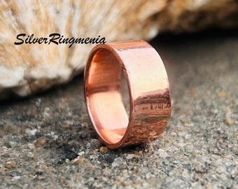 Puede incluir: Un anillo de cobre con una banda ancha, sobre una superficie gris texturizada. La superficie del anillo refleja la luz, mostrando su brillo metálico. El texto "Silver Ringmenia" es visible.