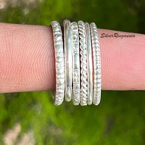 May include: A collection of silver rings with various textures, including smooth, twisted, and patterned designs. The rings are stacked on a finger, showcasing their different styles. The background is a blurred green.