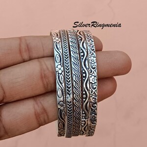 Puede incluir: Un conjunto de cinco pulseras de plata con intrincados diseños florales y geométricos. Las pulseras se sostienen en una mano, mostrando los diseños detallados. El texto "Silver Ringmenia" es visible en la esquina superior derecha.