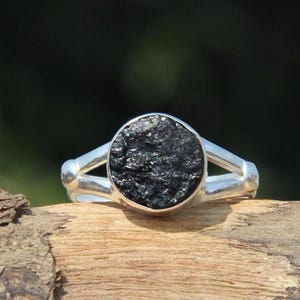 May include: A silver ring featuring a circular, rough-textured black stone set in the centre. The band of the ring splits into two distinct silver arms that connect to the sides of the stone setting, creating a polished metallic finish.