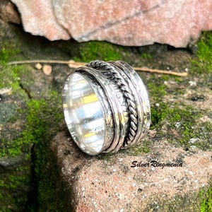 Può includere: Un anello d'argento con un design strutturato. L'anello presenta una fascia intrecciata e più fasce strutturate. L'anello è posto su uno sfondo di muschio verde e una roccia marrone-rossastra. Il testo "Silver Ringmenia" è visibile.