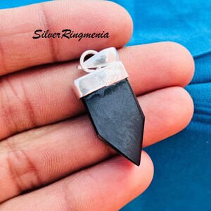 Peut inclure: Un pendentif en pierre noire avec une bélière argentée. Le pendentif est une pierre sombre et polie de forme géométrique. Le haut du pendentif est serti dans un métal argenté. Le texte "Silver Ringmenia" est visible en haut à gauche.