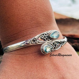 Könnte beinhalten: Ein silbernes Armreif-Armband mit zwei tropfenförmigen, hellblauen Edelsteinen. Das Armband hat verzierte Schnörkel und kleine Perlenakzente. Der Text "Silver Ringmenia" ist sichtbar.