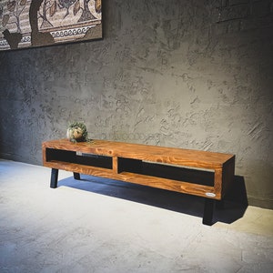 May include: A wooden TV stand with a warm brown finish and black legs. The stand features two open compartments and a small plant on top. The design is modern and minimalist, suitable for a living room.