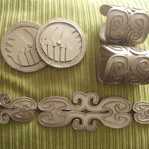May include: A set of four gold-colored metal costume jewelry pieces. Two round pieces with a swirl design, two bracelets with a swirl design, and a belt with a swirl design.