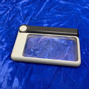 May include: A rectangular magnifying glass with a gray frame and a black handle. The lens is clear, and the handle has a white circular button. The magnifying glass is on a blue velvet surface.