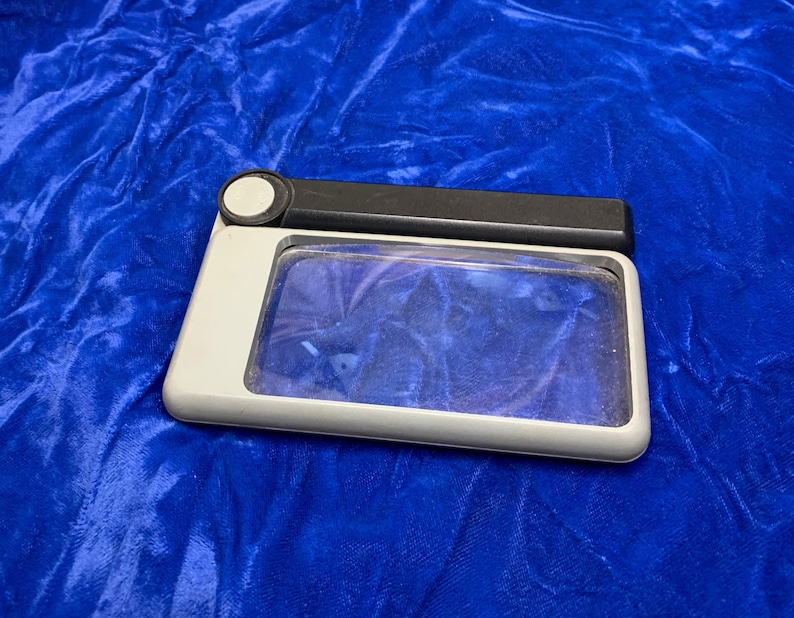 May include: A rectangular magnifying glass with a gray frame and a black handle. The lens is clear, and the handle has a white circular button. The magnifying glass is on a blue velvet surface.