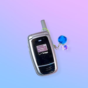 Peut inclure: Téléphone portable argenté à clapet avec un écran noir affichant "Searching for service..." et le logo Verizon. Un porte-clés pompon bleu avec un cristal en forme d'étoile est suspendu au téléphone. Le fond est un dégradé de bleu et de violet.