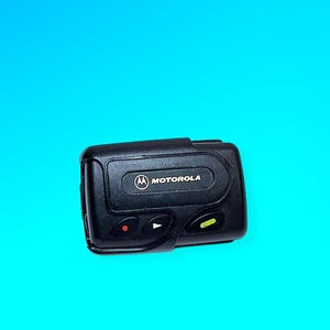 May include: A black Motorola pager with a rectangular design. The pager has a screen and several buttons, including a red and green indicator light. The Motorola logo is visible on the front.
