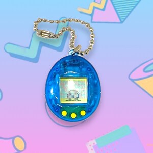 May include: A blue handheld digital pet game with a silver chain. The game has a small screen displaying pixelated graphics and three yellow buttons below. The device is a nostalgic toy from the 90s.