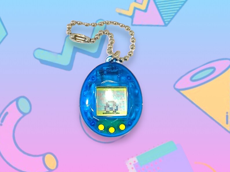 May include: A blue handheld digital pet game with a silver chain. The game has a small screen displaying pixelated graphics and three yellow buttons below. The device is a nostalgic toy from the 90s.