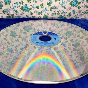 May include: A clear, circular disc with a floral pattern overlay. The disc has a rainbow reflection and a blue label that reads "DISCOVISION" and "BLUE COLLAR 1". The background is a blue surface and a floral fabric.
