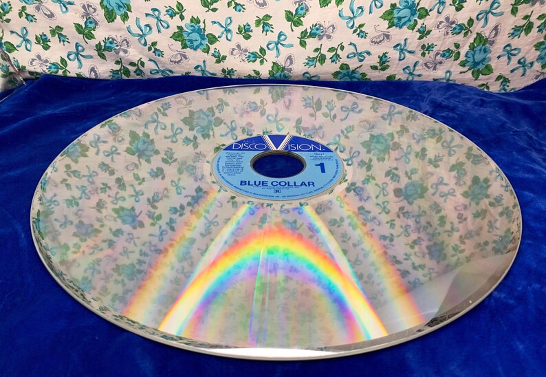 May include: A clear, circular disc with a floral pattern overlay. The disc has a rainbow reflection and a blue label that reads "DISCOVISION" and "BLUE COLLAR 1". The background is a blue surface and a floral fabric.