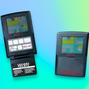 Op de afbeelding: Twee vintage draagbare elektronische spellen. Eén is open en toont een blackjack-spel met de tekst "CAESARS PALACE" en "HOUSE RULES". De andere is gesloten, met de tekst "TIGER" en "CAESARS PALACE" in het rood.