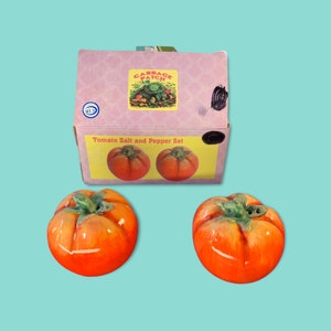 Puede incluir: Un juego de salero y pimentero con forma de tomate con una caja a juego. Los saleros y pimenteros de cerámica son de color naranja con detalles de hojas verdes. La caja es rosa con una etiqueta amarilla que dice "Tomato Salt and Pepper Set".