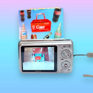 May include: A silver Sony digital camera with a wrist strap, displaying an image of a miniature Coca-Cola carrier and bottles. A colorful background features a gradient of blue, pink, and purple.
