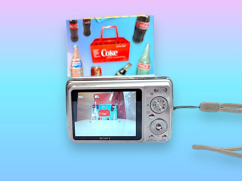 May include: A silver Sony digital camera with a wrist strap, displaying an image of a miniature Coca-Cola carrier and bottles. A colorful background features a gradient of blue, pink, and purple.