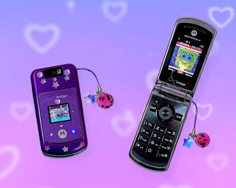May include: Two Motorola flip phones, one purple and one black, with screens displaying images. The purple phone has star and gem embellishments. Both phones have charms with pink and black pom-poms and blue star accents. The background is a gradient of purple and pink with white heart shapes.