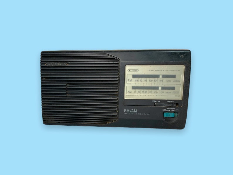 Może przedstawiać: Czarny radioodbiornik FM/AM Sony z pokrętłem strojenia i wyświetlaczem częstotliwości. Radio posiada przycisk zasilania i selektor pasma. Na panelu radia widnieje napis "FM/AM" i "SONY".