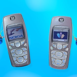 Peut inclure: Deux téléphones portables Nokia argentés avec des accents bleus et des breloques. Les écrans affichent des images et le logo Nokia. Les téléphones ont un clavier avec des chiffres et des boutons de fonction. Le fond est bleu uni.