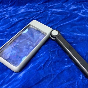 May include: A rectangular magnifying glass with a gray frame and a black handle. The handle is folded out, and the lens is clear. The magnifying glass is set against a blue background.