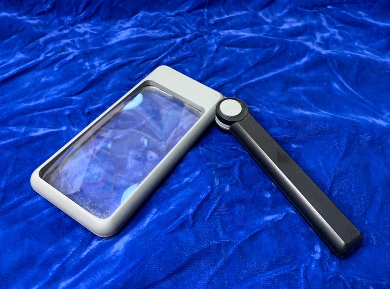 May include: A rectangular magnifying glass with a gray frame and a black handle. The handle is folded out, and the lens is clear. The magnifying glass is set against a blue background.