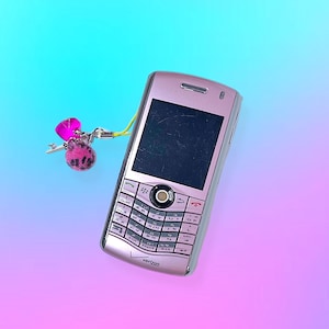 Peut inclure: Un téléphone portable rose avec un écran noir et un clavier. Un porte-clés rose et noir avec une clé et une gemme rose est attaché au téléphone avec une lanière jaune. Le téléphone porte le logo Verizon.