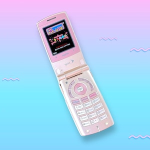 Puede incluir: Un teléfono móvil rosa Sanyo con un juego de Pac-Man en la pantalla. El teléfono está abierto, mostrando el teclado y la pantalla. El fondo es un degradado de azul y rosa con formas de onda.