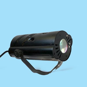 Peut inclure: Un projecteur de scène noir avec une lentille et une poignée. La lumière a un corps cylindrique avec des fentes de ventilation et un cordon d'alimentation. La lentille est entourée d'un cadre décoratif festonné. La lumière est posée sur un fond bleu clair.